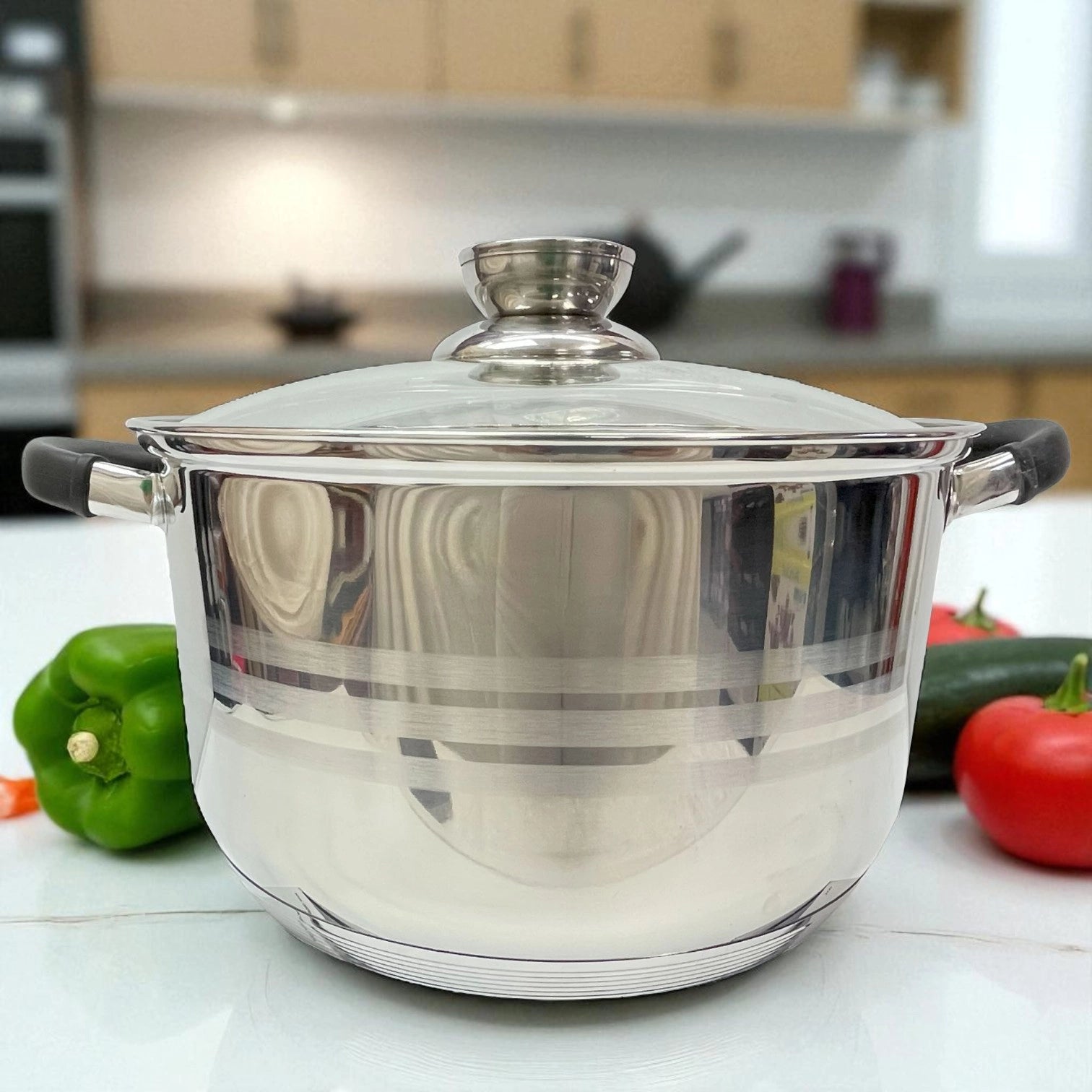 Olla de acero inoxidable MGFR con tapa de vidrio y asas de silicona negra, vista lateral en ambiente moderno de cocina, decorada con tomate y pimiento verde.