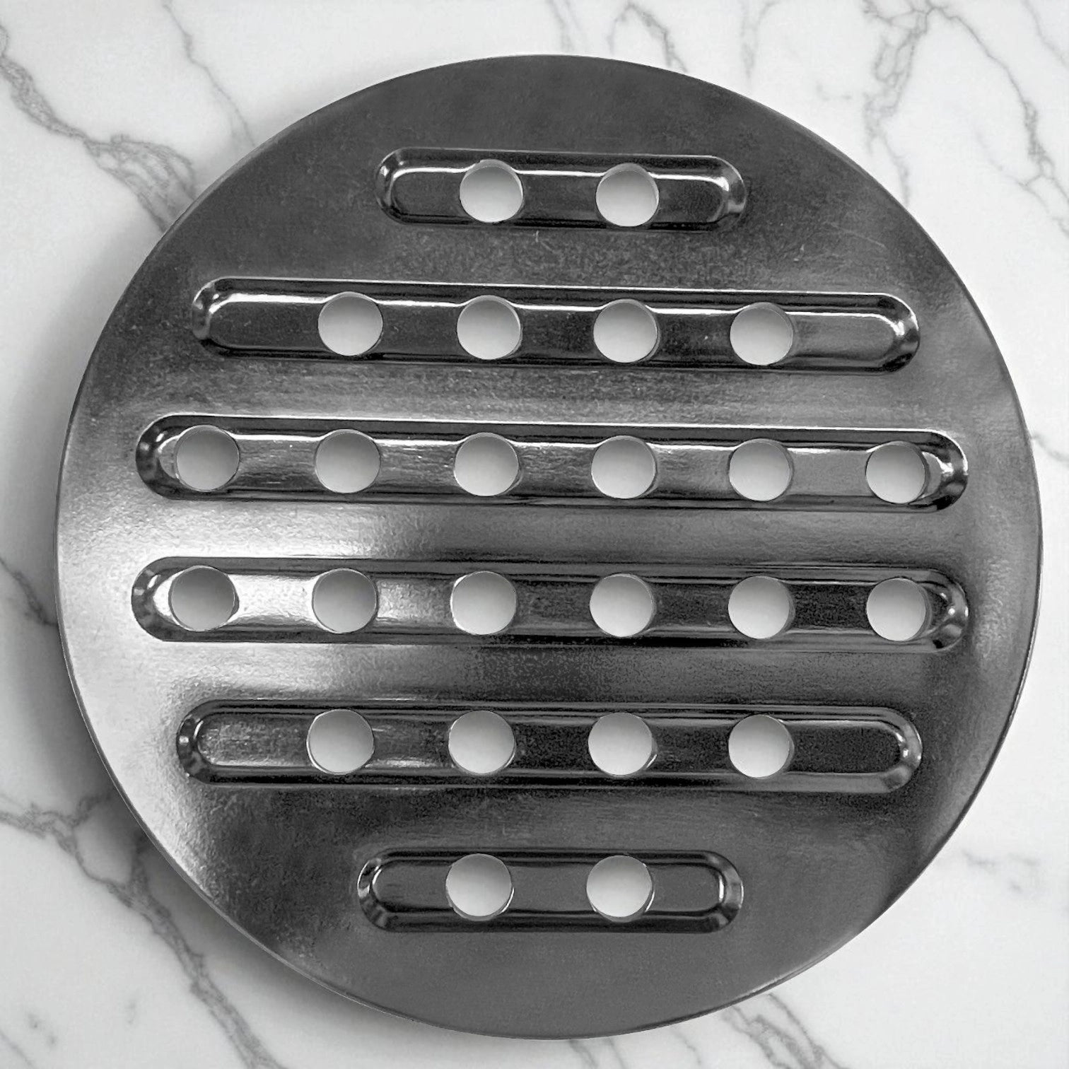 Difusor de calor circular de acero inoxidable con perforaciones, ideal para estufa de gas o vitrocerámica, sobre encimera de mármol blanca.