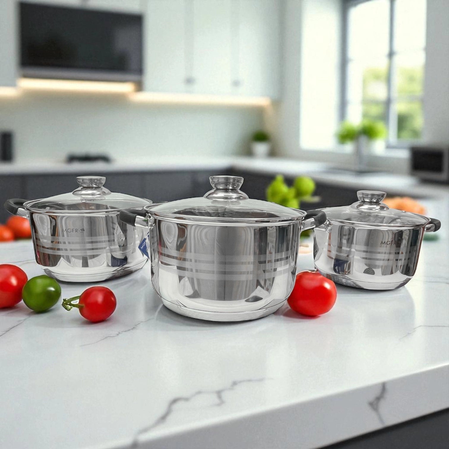 Trío de ollas MGFR de acero inoxidable 18/10 con tapas transparentes, asas negras anticalóricas, colocadas sobre encimera de mármol blanco con tomates y vegetales como decoración.