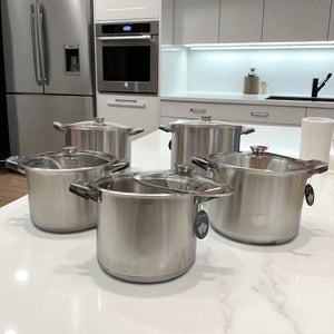 Set de 10 piezas de ollas Epicure de acero inoxidable con tapas de vidrio, distribuidas sobre isla blanca en cocina minimalista.