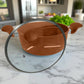 Olla mediana color bronce con tapa de vidrio semiabierta, vista diagonal en cocina moderna.