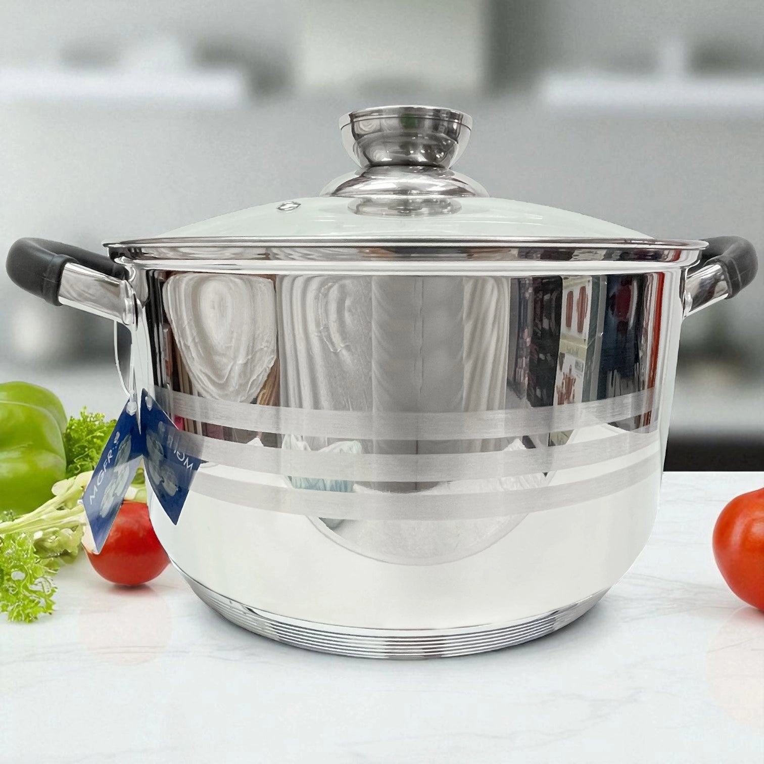 lla de acero inoxidable MGFR con tapa de vidrio templado, asas de silicona negras y diseño de líneas satinado, sobre encimera de mármol con tomates decorativos al costado.