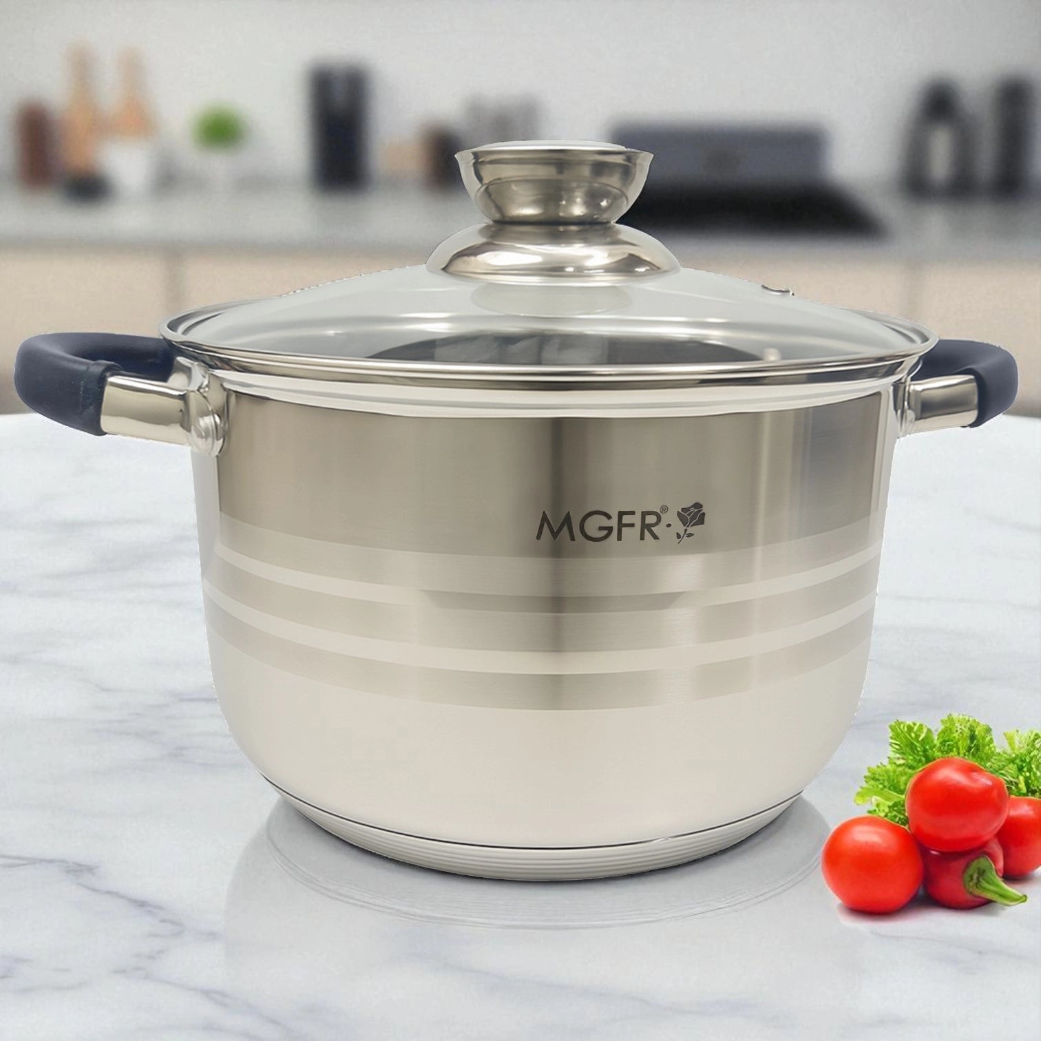 Olla de acero inoxidable MGFR con tapa de vidrio templado, asas de silicona negras y diseño de líneas satinado, sobre encimera de mármol con tomates decorativos al costado