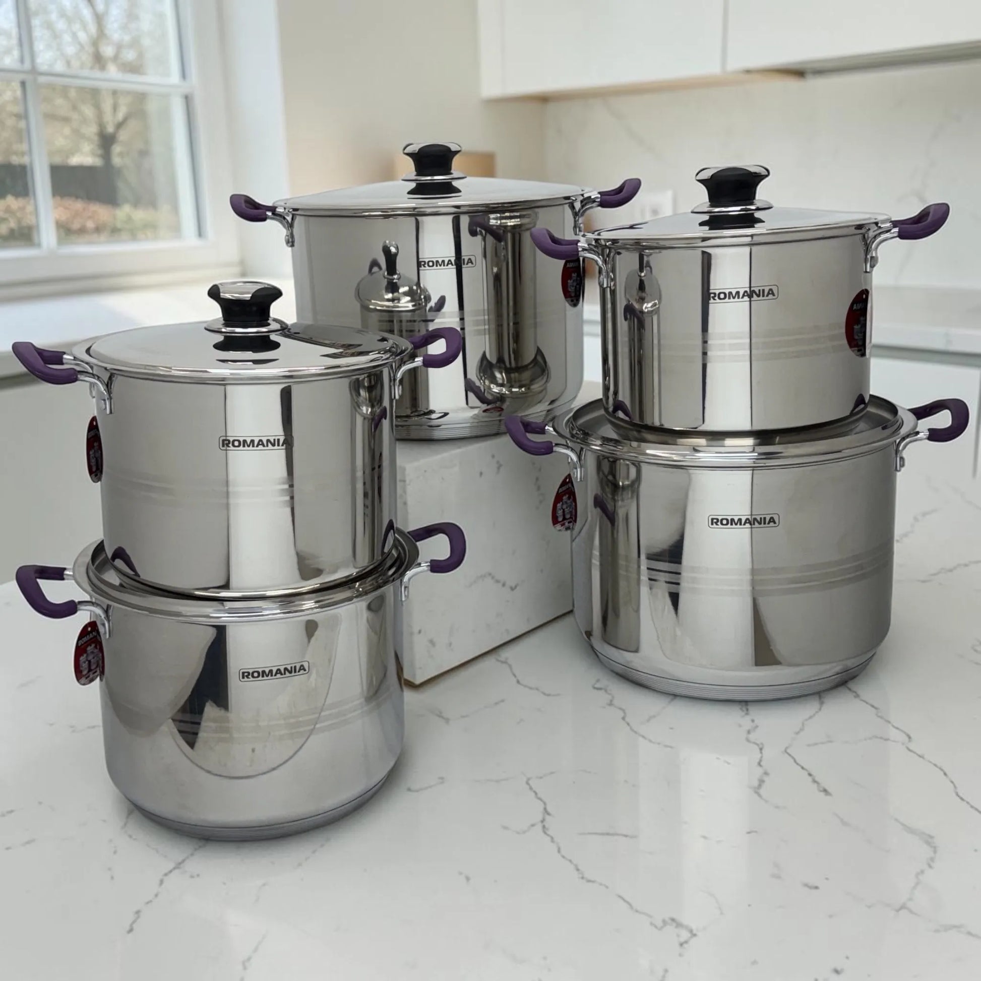 Batería de cocina Romania de 10 piezas en acero inoxidable con mangos púrpura, colocada sobre encimera blanca en cocina moderna.