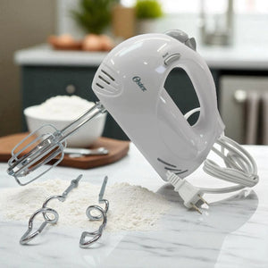 Batidora Oster blanca de mano con cable y batidores instalados, acompañada de ganchos amasadores sobre mesa de cocina.