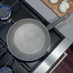 Vista superior de sartén Bergner Orion Ø32 cm gris con antiadherente Marble+ sobre estufa de gas; escena de cocina con huevos al fondo.