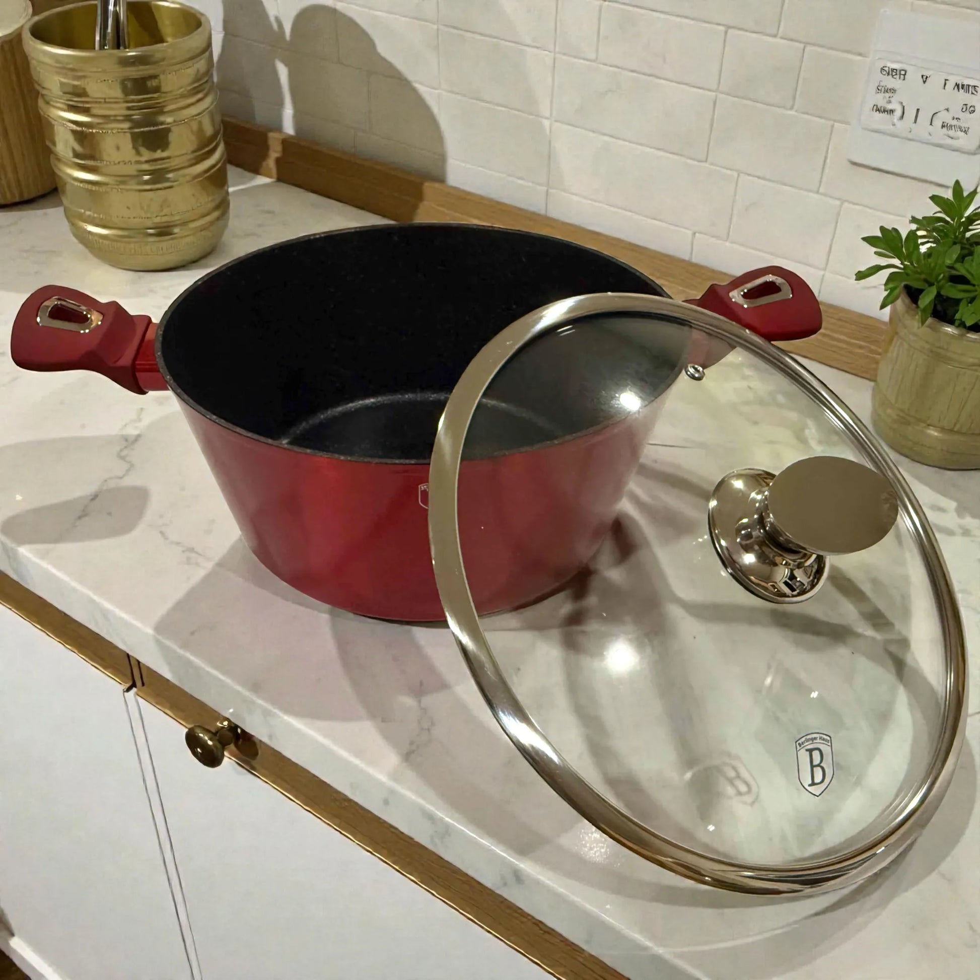 Detalle de cacerola Berlinger Haus Metallic Line Burgundy con tapa de vidrio y pomo metalico, vista en angulo sobre encimera, interior antiadherente tipo marmol.