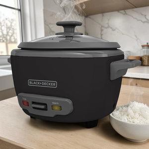 Olla arrocera BLACK+DECKER RC514 de 14 tazas cocidas, color negro con tapa de vidrio, cocinando con vapor sobre mesa de cocina junto a un tazón de arroz.