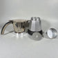 Componentes de cafetera moka eléctrica MOKA 3–6 tazas en fondo claro: jarra superior con asa, caldera inferior plateada, filtro/placa y embudo.