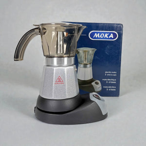 Cafetera moka eléctrica MOKA 3–6 tazas ensamblada sobre la base, vista frontal, con caja del producto al fondo; acabados plata y negro.