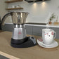 Cafetera moka eléctrica MOKA 3–6 tazas sobre base calefactora junto a taza y plato; escena de cocina moderna, colores plata, negro y gris.