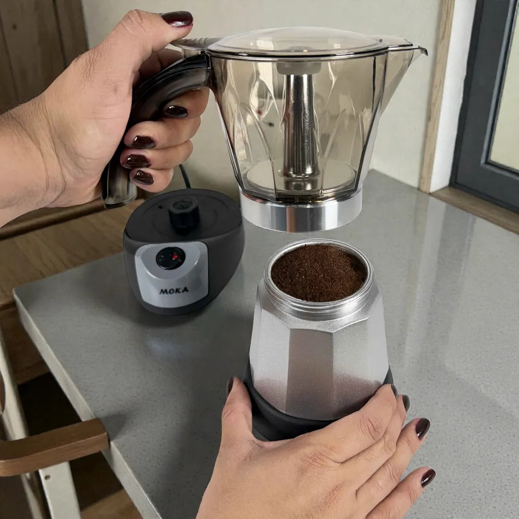 Manos desmontando la cafetera moka eléctrica MOKA 3–6 tazas: jarra superior separada y caldera inferior con café molido; base calefactora al fondo.