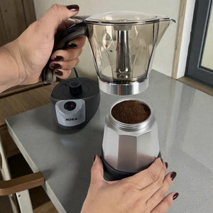 Manos desmontando la cafetera moka eléctrica MOKA 3–6 tazas: jarra superior separada y caldera inferior con café molido; base calefactora al fondo.