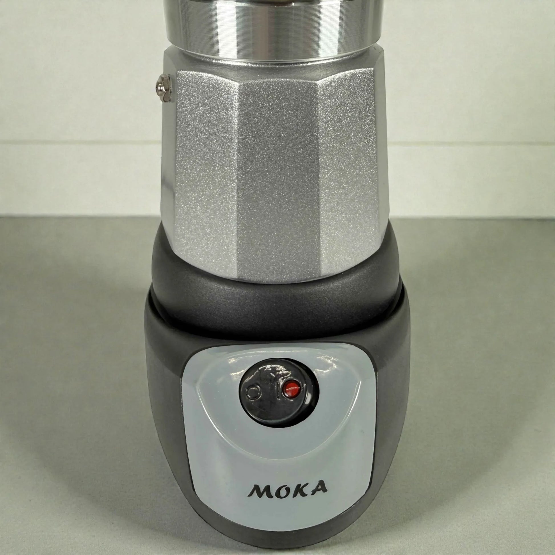 Detalle de la cafetera moka eléctrica MOKA 3–6 tazas: base calefactora con interruptor e indicador luminoso, cuerpo plateado y panel frontal con marca MOKA.