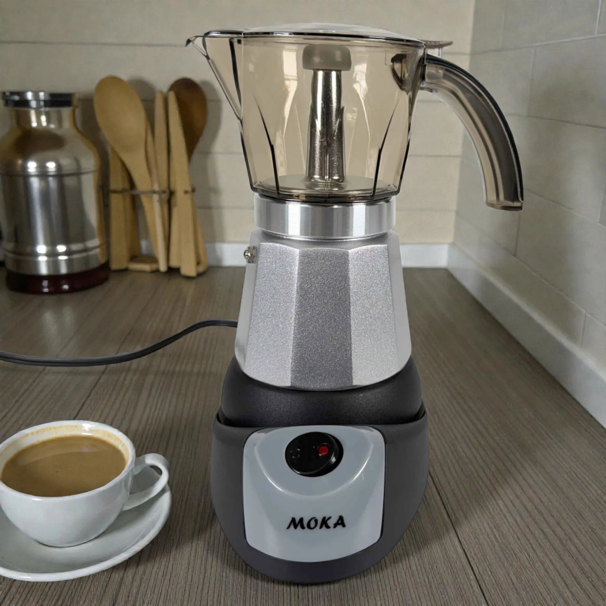 Cafetera moka eléctrica MOKA 3–6 tazas ensamblada sobre base con logo MOKA; taza de café al lado en encimera, acabado plata y negro.