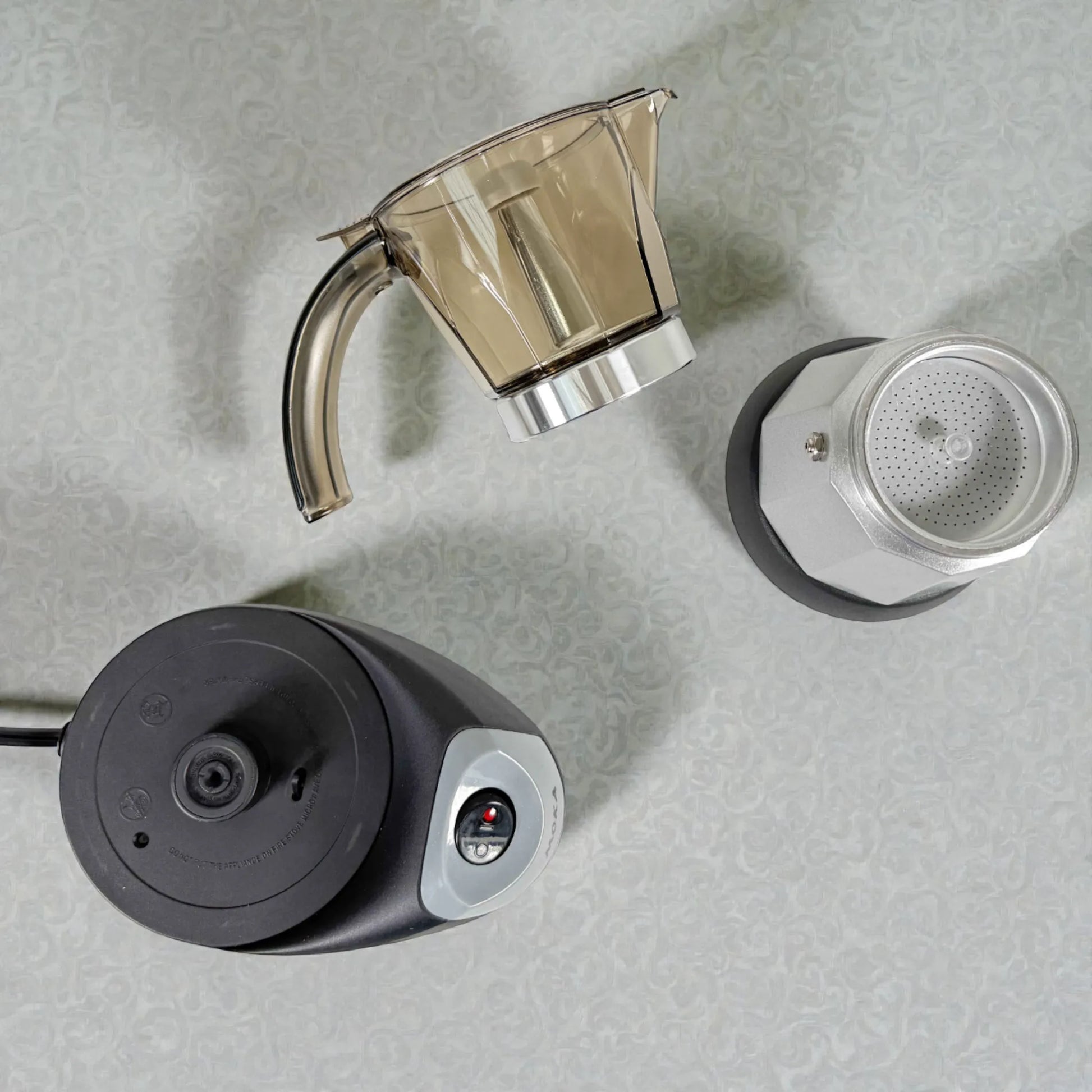 Vista superior de cafetera moka eléctrica MOKA 3–6 tazas desarmada: base calefactora, jarra superior y caldera inferior plateada.