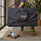 Persona cargando en el hombro la bolsa de transporte negra de la cama de masaje plegable Lotus Home Collection, demostrando su diseño portátil y fácil de mover.