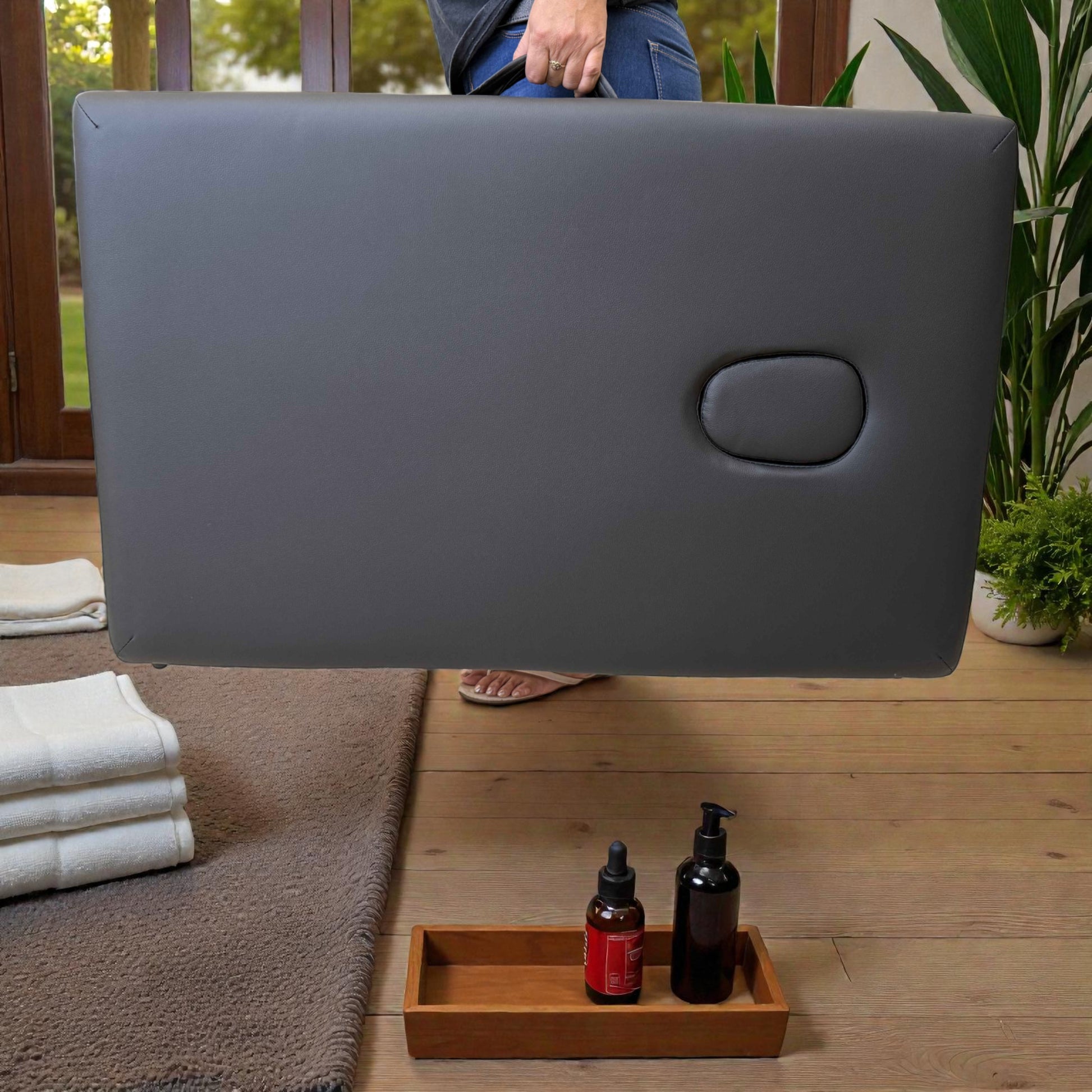 Persona cargando cama de masaje plegable Lotus Home Collection color negro, completamente plegada tipo maleta de 92x60x10 cm, mostrando superficie acolchada y tapón del orificio facial.