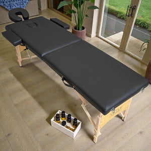 Cama de masaje plegable Lotus Home Collection color negro, vista diagonal en salón de spa, con patas de madera clara, reposacabezas ajustable y apoyabrazos laterales.