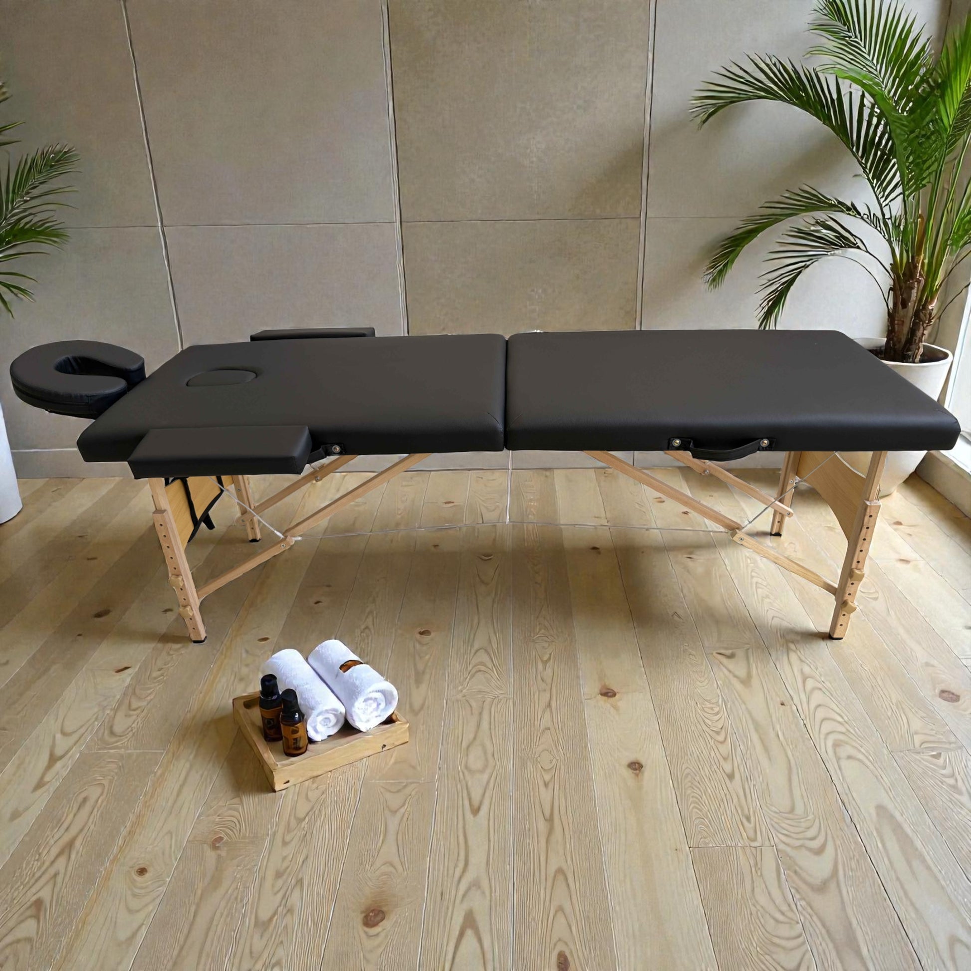 Cama de masaje negra Lotus plegable y portátil, vista lateral completa, con patas regulables en altura y accesorios de spa como toallas y aceites en el suelo.