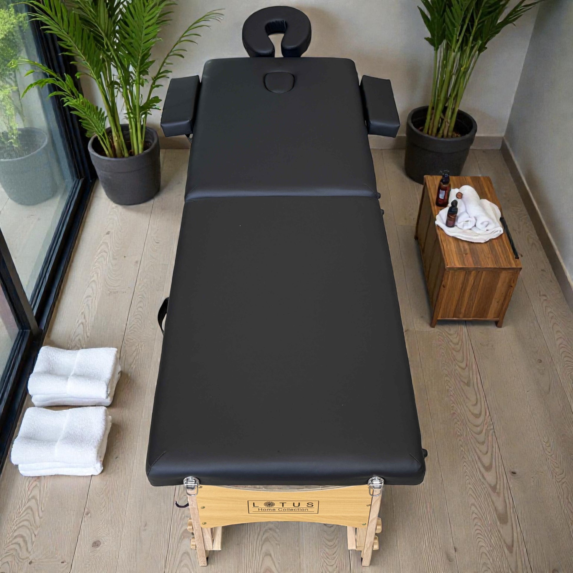 Vista superior de cama de masaje plegable Lotus negra de 216x80 cm, totalmente extendida, con apoyabrazos laterales, reposacabezas y accesorios de spa como toallas y aceites.