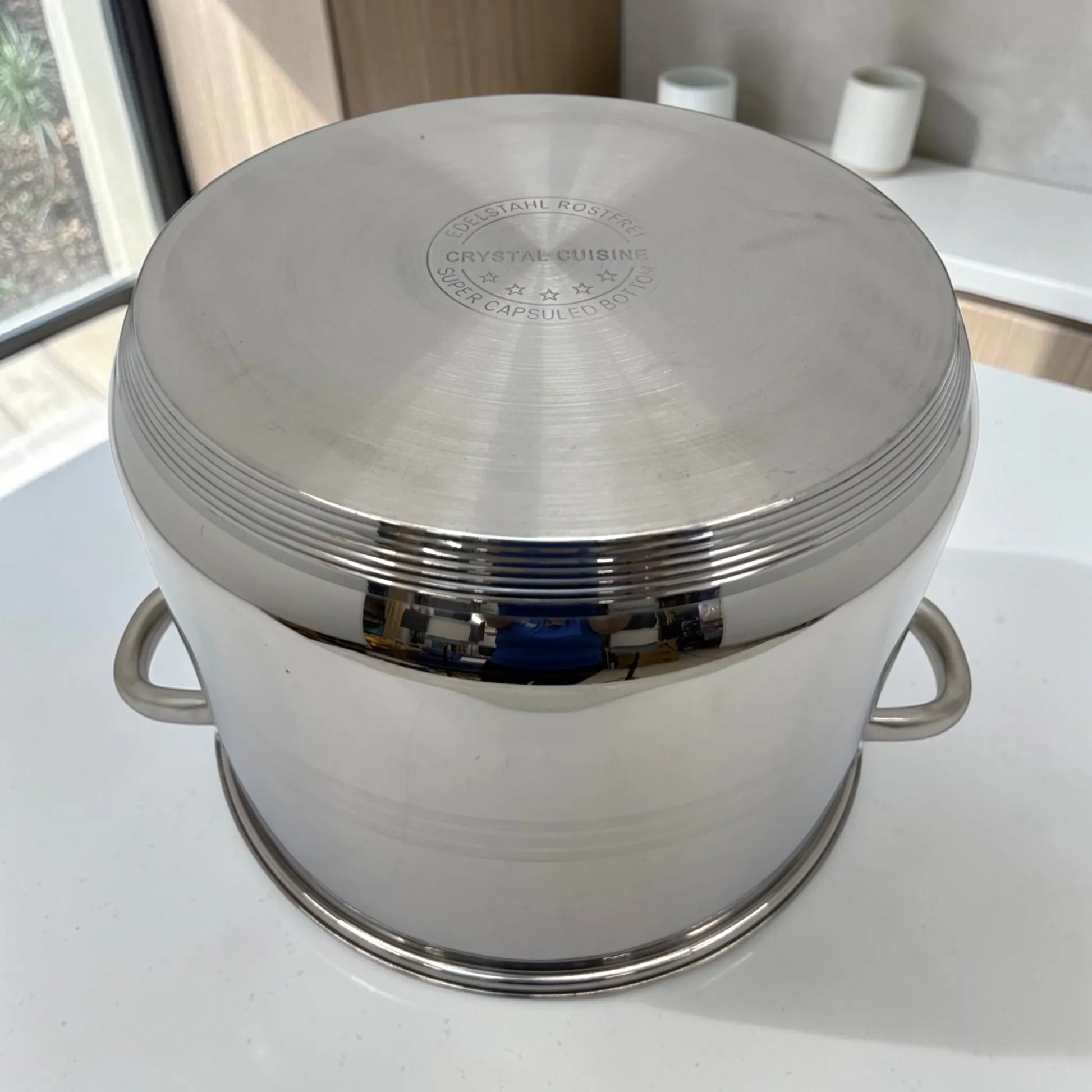 Olla Crystal Cuisine CC-138 de acero inoxidable color plateado, colocada boca abajo para mostrar la base encapsulada y el fondo multicapa sobre encimera blanca.