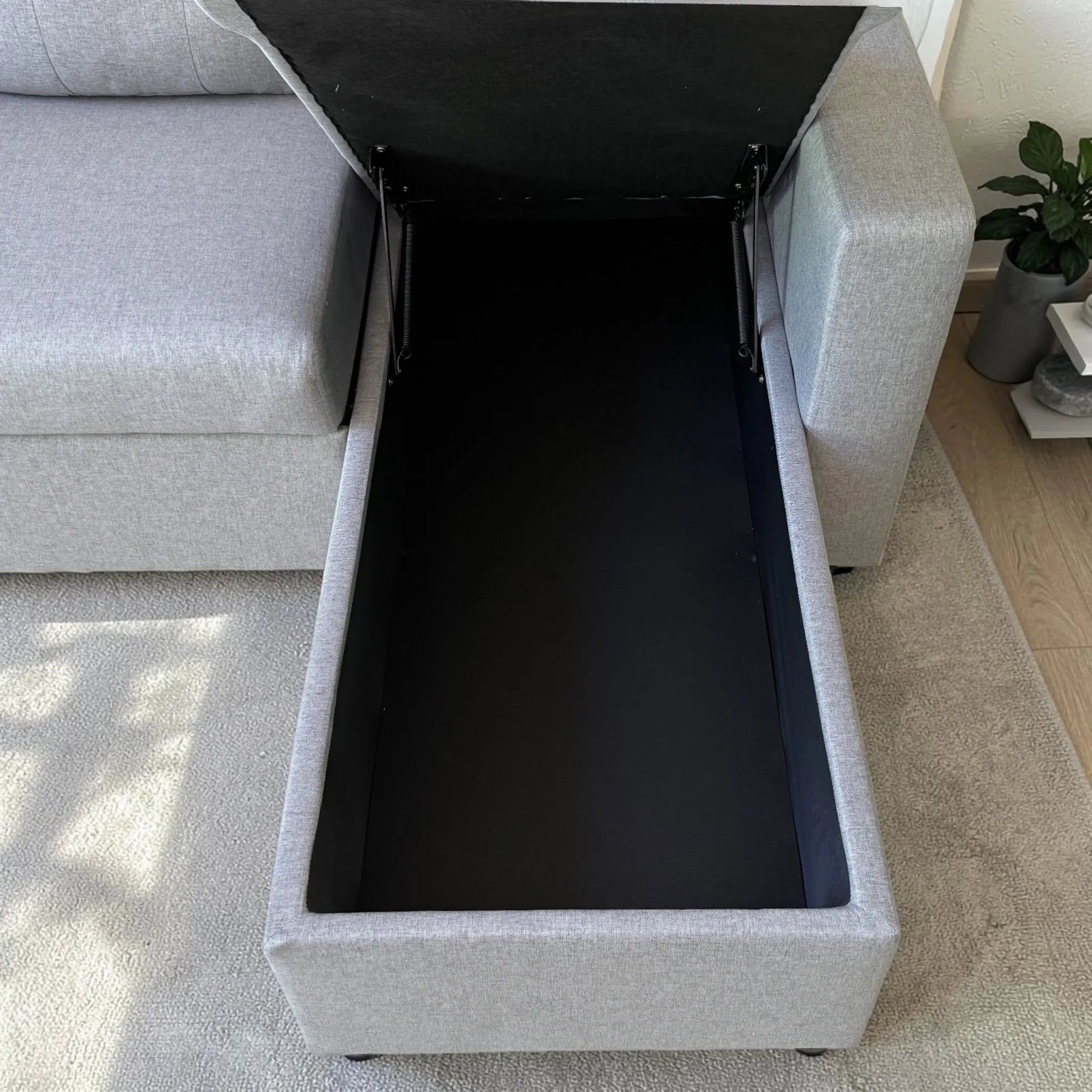 Detalle del cajón de almacenamiento del sofá cama gris claro Ambienta Home Decor, interior negro amplio, tapa levantada y estructura lateral tapizada.