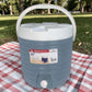 Dispensador térmico Princeware gris de 17 litros SKU 3554 con asa blanca, tapa superior y grifo frontal, colocado sobre mantel de picnic al aire libre.