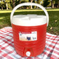 Dispensador térmico Princeware rojo de 17 litros SKU 3554 con asa blanca, tapa superior y grifo frontal, colocado sobre mantel de picnic al aire libre.