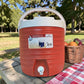 Dispensador térmico Princeware rojo de 17 litros SKU 3554 con asa, tapa blanca, grifo frontal y canasta de frutas en ambiente de picnic.