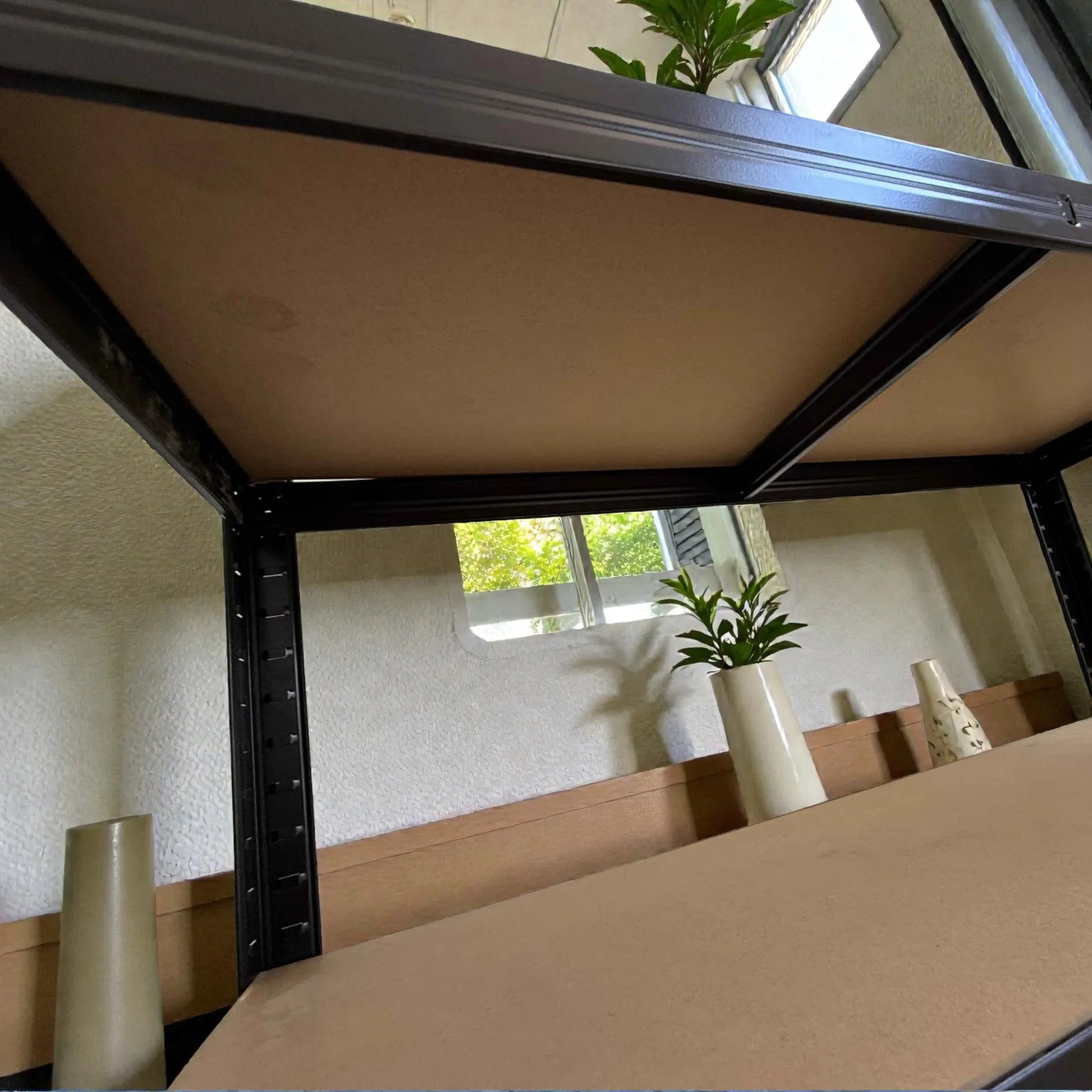 Vista desde abajo del estante anaquel metálico Lotus de 5 niveles 90x40x180 cm, mostrando refuerzos metálicos y parte inferior de las repisas MDF con plantas al fondo.