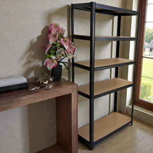 Vista lateral del estante anaquel metálico Lotus de 5 niveles 90x40x180 cm, estructura negra con repisas MDF, colocado en interior junto a mesa de madera y ventana.