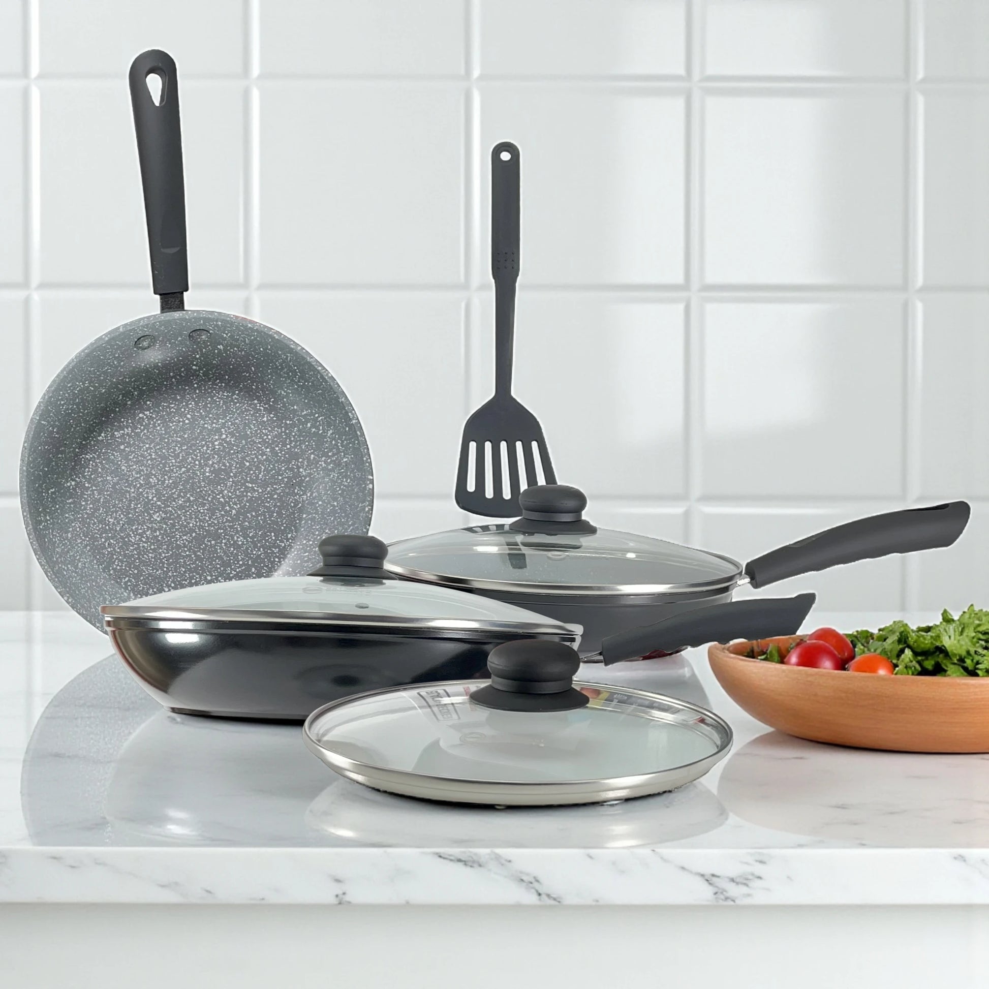 Set de sartenes Estrella Gourmet interior gris marmolizado y exterior negro, con tapas de vidrio y espátula de nylon sobre encimera de mármol.