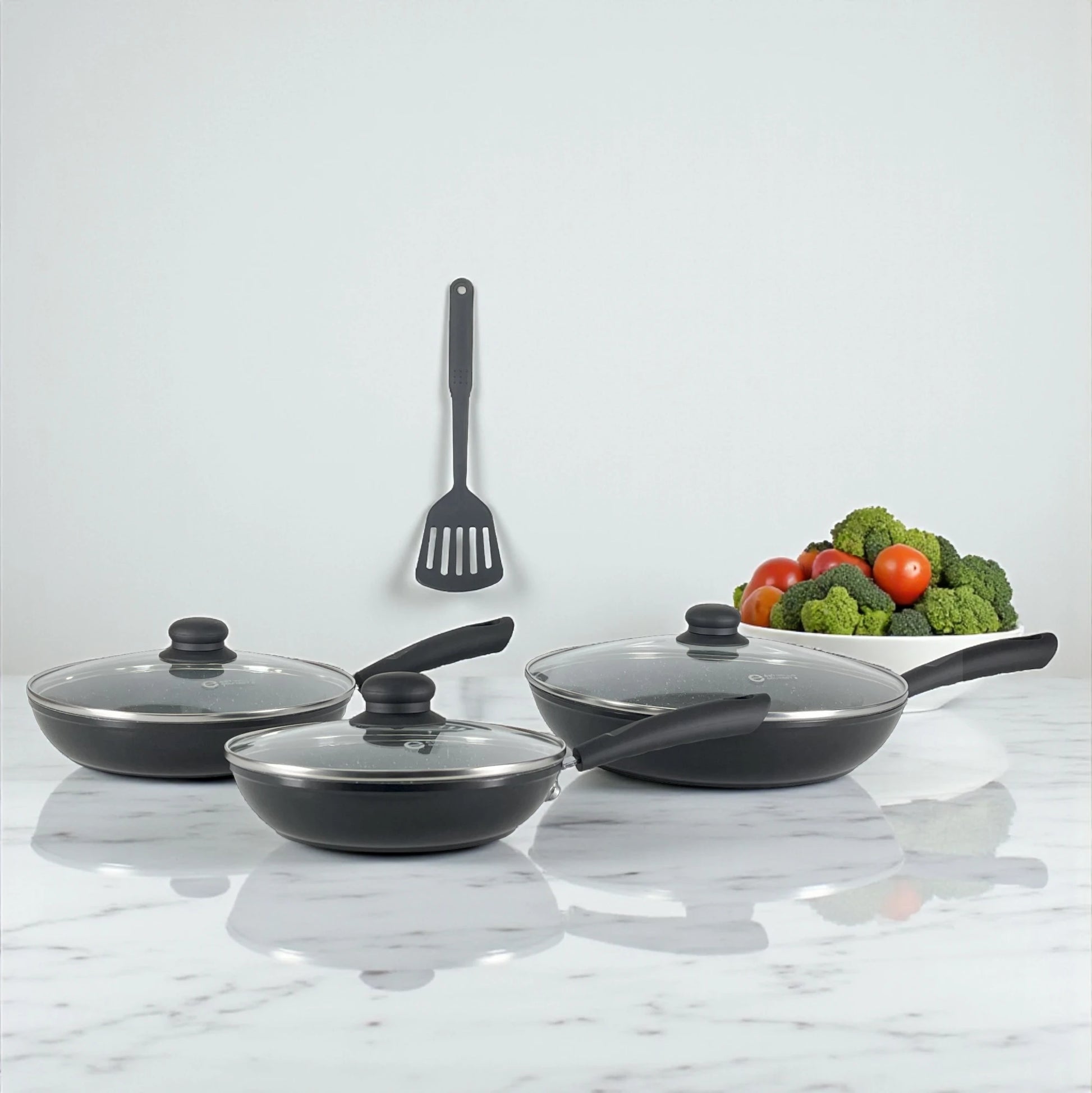 Tres sartenes Estrella Gourmet con tapas de vidrio y espátula colgada, acabado exterior negro e interior gris marmolizado.