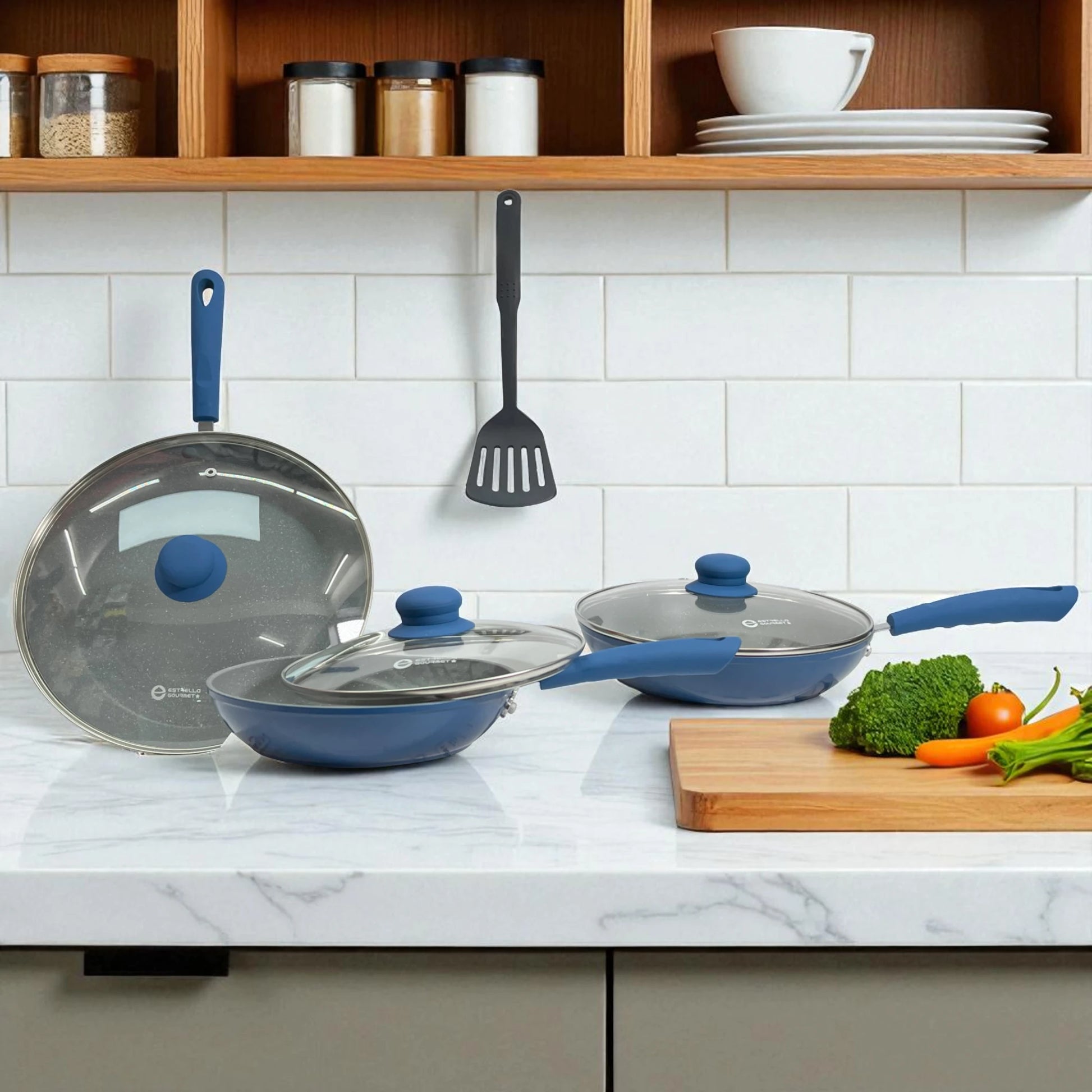 Tres sartenes Estrella Gourmet azules con tapas de vidrio y mangos de baquelita sobre encimera; espátula colgada al fondo; set 20, 24 y 30 cm.
