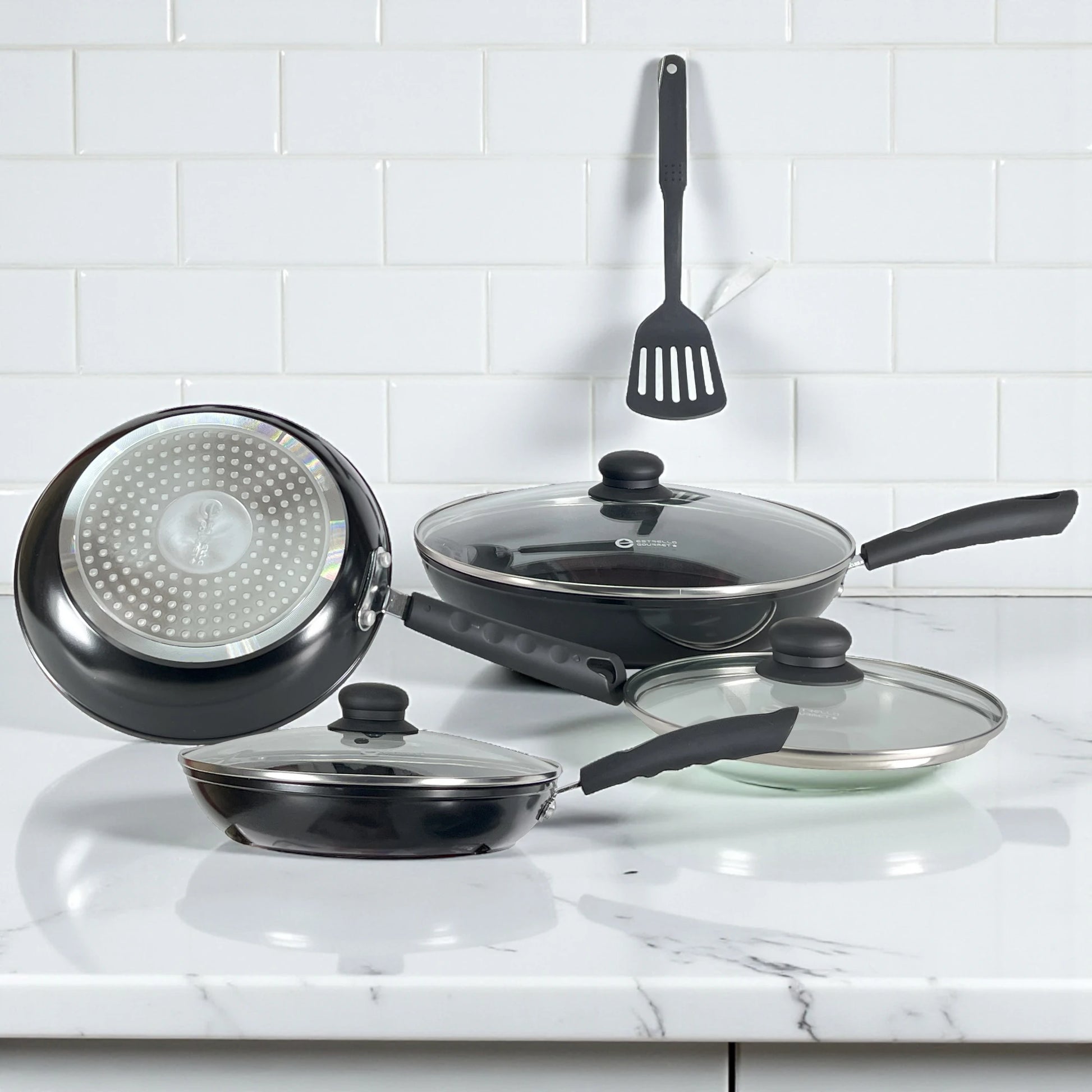 Primer plano de la base de acero difusor para inducción del sartén Estrella Gourmet; junto a sartenes con tapa, acabado negro en aluminio forjado.