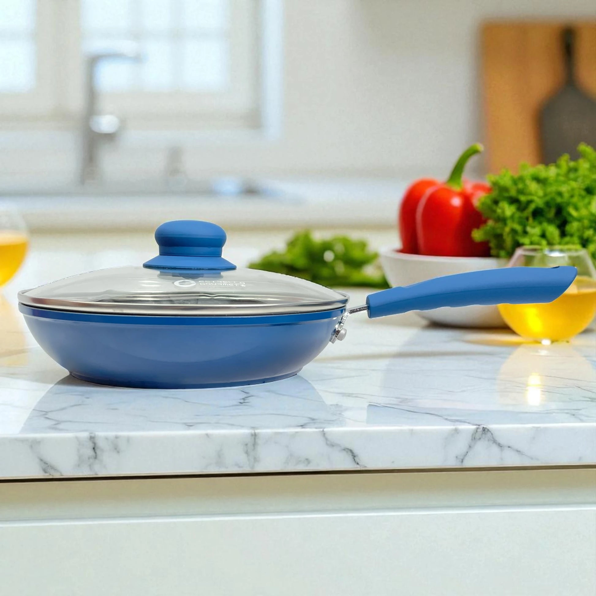 Sartén Estrella Gourmet color azul con tapa de vidrio templado y mango ergonómico de baquelita, vista lateral sobre encimera; línea marmolizada disponible en 20, 24 y 30 cm.