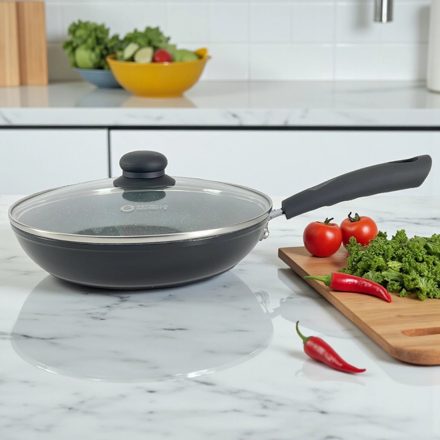 Sartén Estrella Gourmet con tapa de vidrio, interior gris marmolizado y mango de baquelita a la derecha sobre encimera de mármol; verduras y chiles a un lado, tamaños disponibles 20, 24 y 30 cm.