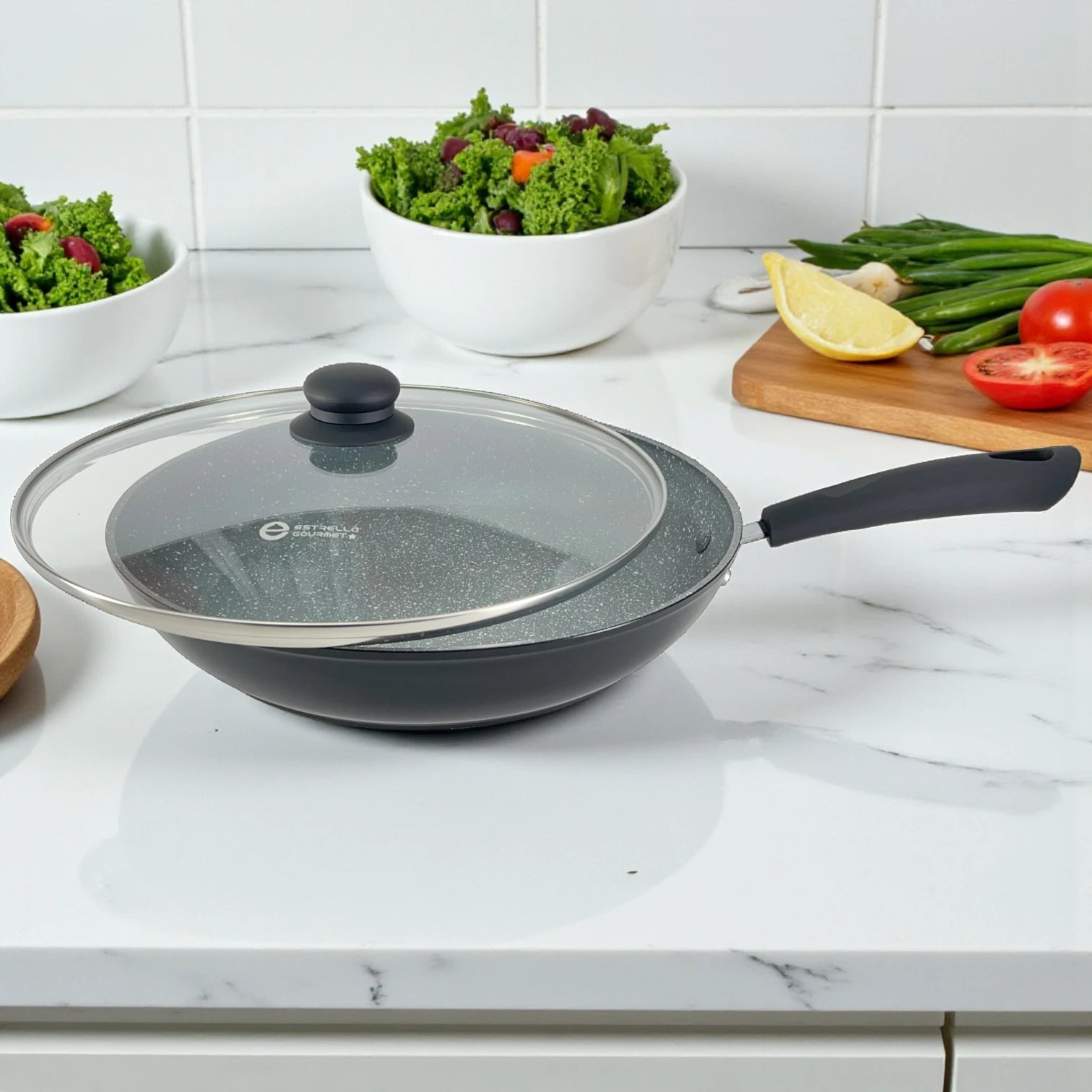 Sartén Estrella Gourmet con recubrimiento marmolizado y tapa de vidrio semiabierta, interior gris y mango de baquelita; bowls de ensalada al fondo, tamaños 20, 24 y 30 cm.