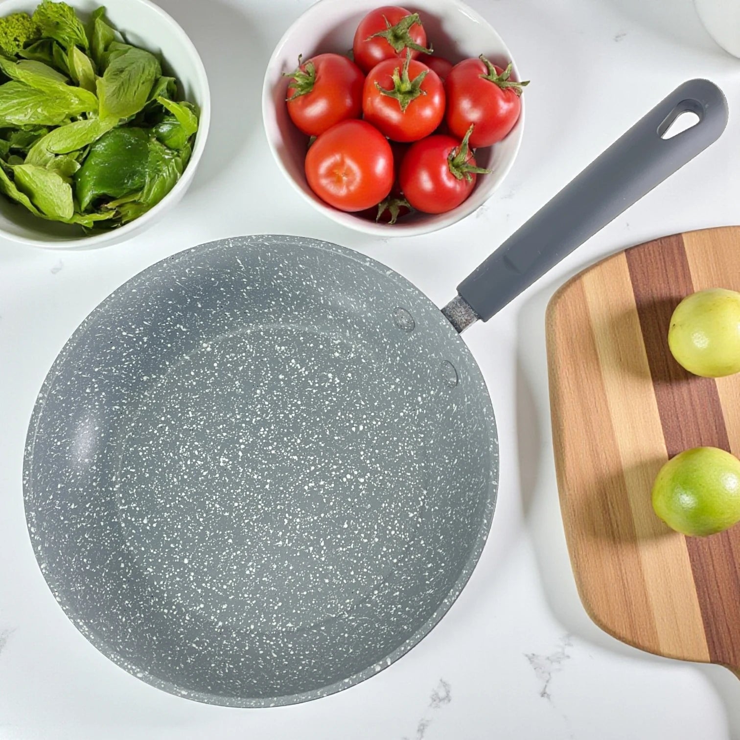 Vista superior del sartén Estrella Gourmet con acabado tipo piedra, interior marmolizado gris y mango de baquelita; cuencos con tomates y hierbas alrededor; tamaños 20, 24 y 30 cm.