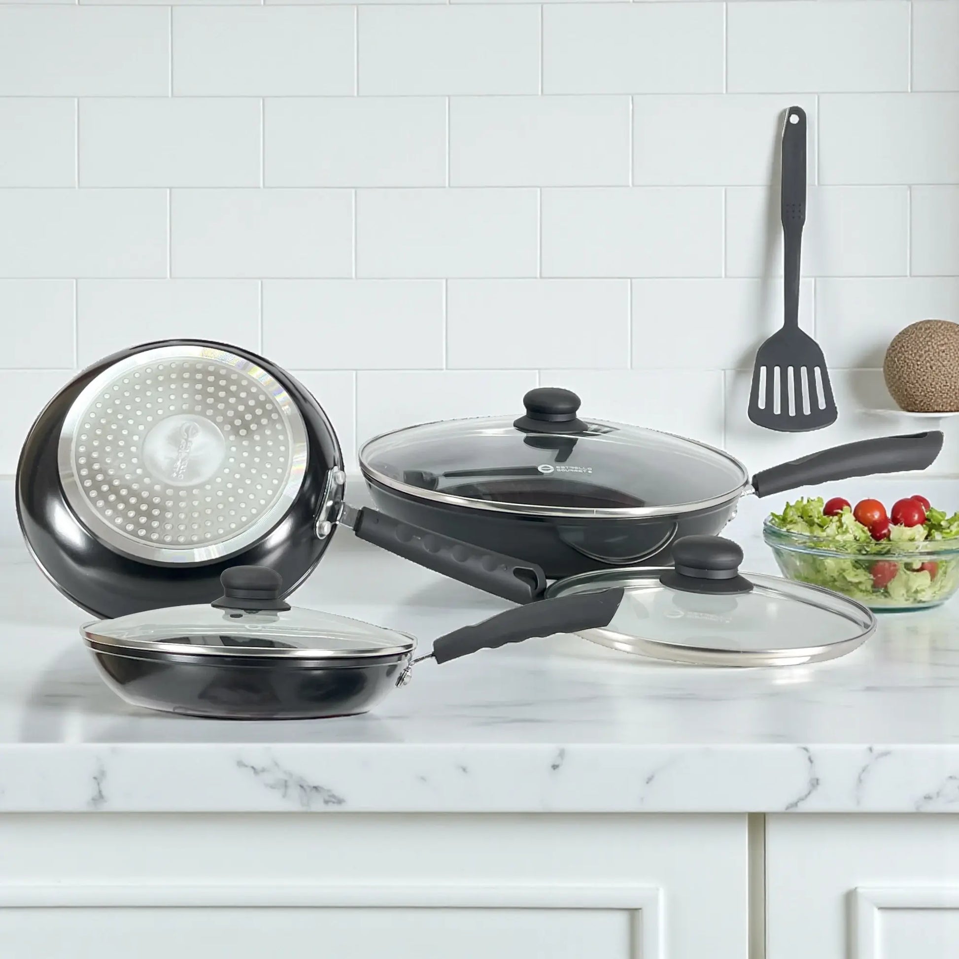 Vista que muestra la base difusora compatible con inducción de un sartén Estrella Gourmet y dos sartenes con tapa; aluminio forjado negro, línea antiadherente.