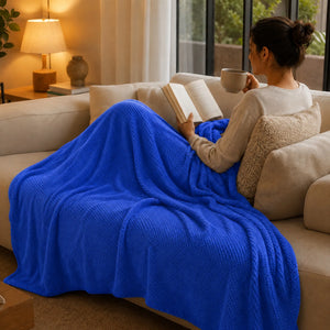 Mujer leyendo y tomando una bebida mientras usa una frazada Colven azul rey de 190 x 190 cm sobre un sofá.