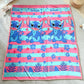 Frazada piel de borrego infantil Stitch con rayas turquesa y rosa, 230x190 cm y 1500 g, tomada extendida en el piso; estampado con Stitch sonriendo, flores y hojas tropicales.