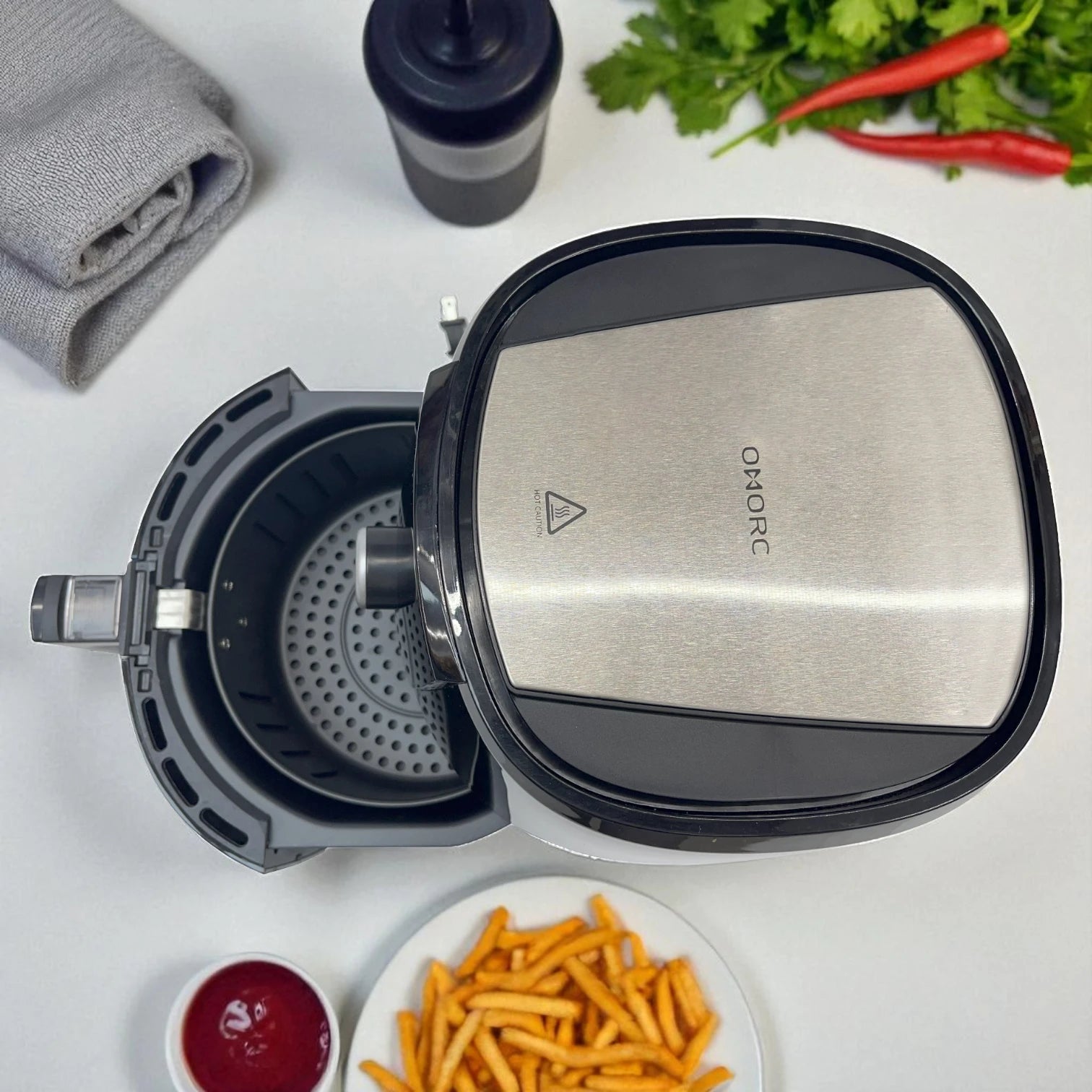 Vista cenital de la freidora de aire OMORC CP176A 4.2 L negra con tapa de acero inoxidable abierta; interior de la cesta con placa perforada, con salsas, chiles y plato de papas en la mesa.