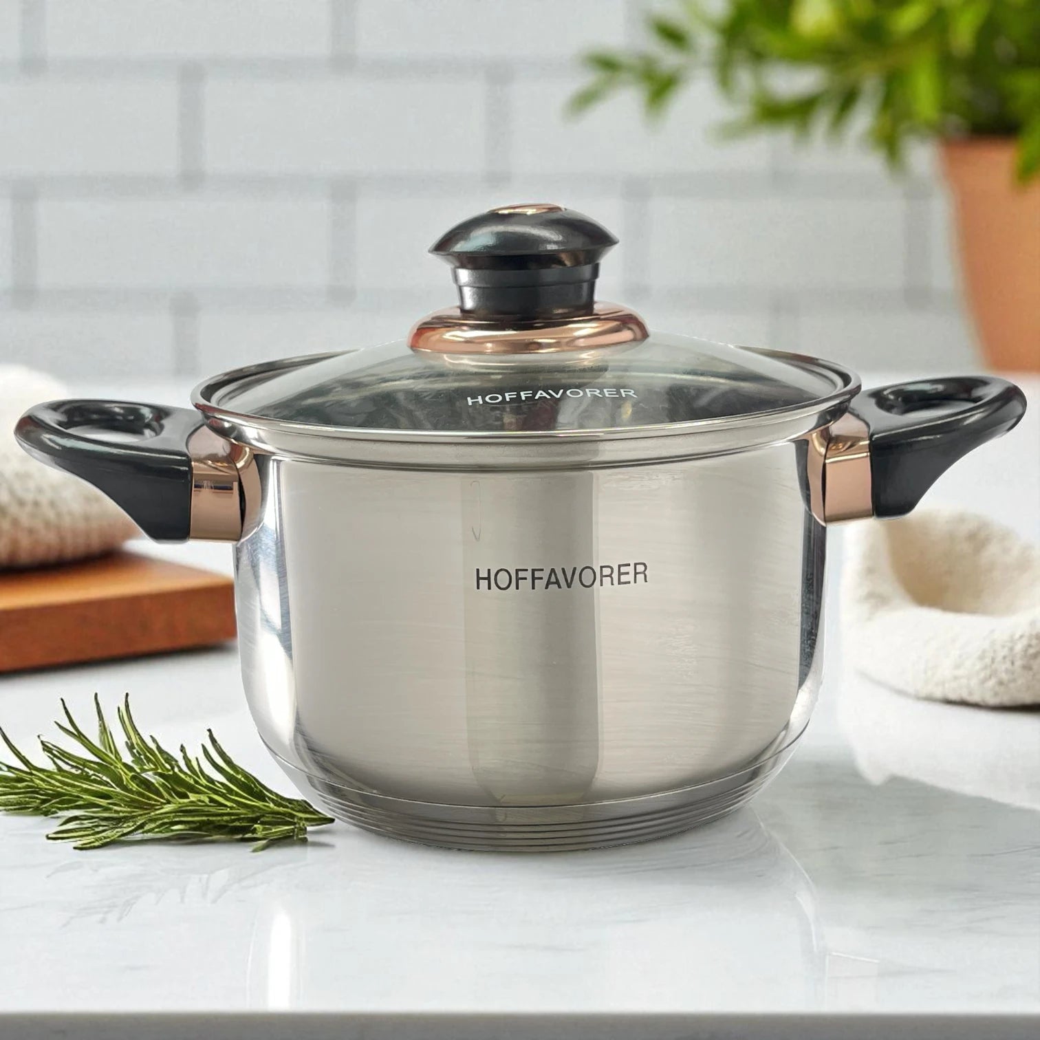 Cacerola pequeña Hoffavorer de 16 cm (2.1 L) en acero inoxidable 18-10, color plateado, con tapa de vidrio y perilla negra con aro dorado, dos asas negras; vista frontal sobre encimera blanca.