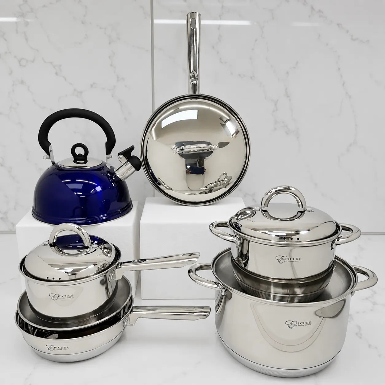 Juego de cocina Epicure by Joseph Marcus con ollas de acero inoxidable, sartén con tapa metálica y tetera azul silbante sobre fondo blanco.