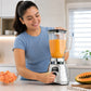 Mujer usando licuadora clásica Oster cromada BLST4655 con jugo de papaya en cocina moderna, imagen lifestyle de producto.