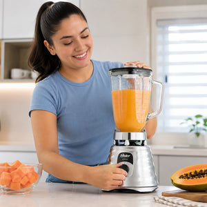 Mujer usando licuadora clásica Oster cromada BLST4655 con jugo de papaya en cocina moderna, imagen lifestyle de producto.