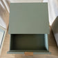 Vista superior de mesa auxiliar STUDIO-1061 verde oliva con cajón abierto, espacio interior amplio y tirador dorado.