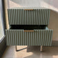 Mesa auxiliar STUDIO-1061 verde oliva con cajón inferior abierto, frente acanalado vertical, tiradores dorados y patas metálicas.